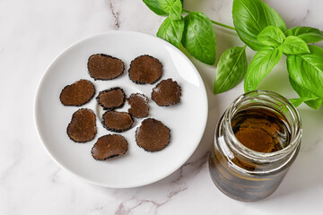 Sliced black truffle on a plate and jar of truffle carpaccio slices in oil over marble background. Canned delicacy mushroom Tuber melanosporum. Gourmet ingredient taste.