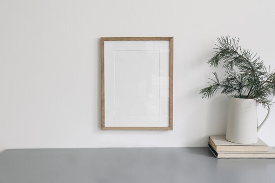 Christmas interior decor. Blank wooden picture frame mockup, white wall. Green pine tree branches in jug and old books. Grey working table. Old books. Winter home office still life. Poster display.