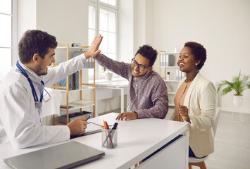 Excited male doctor and ethnic couple give high five celebrate good health results. Smiling surgeon or GP congratulate happy multiethnic family with amazing news. Recovery and medical treatment.