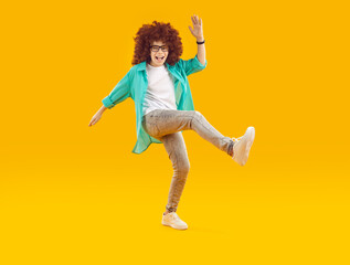 Fototapeta na wymiar Happy cheerful funny boy in red curly wig, dressed in casual clothes, fooling around and dancing in cheerful mood. Full-size isolated shot on orange background.