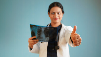 Portrait of a young female doctor examining patient's x-rays scan, Cheerful Asian Indian woman doctor in apron and stethoscope isolated over blue studio background