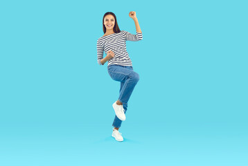 Fototapeta na wymiar Full-size photo of a happy young brunette with straight long hair in a striped jumper and blue jeans rejoices in victory. Yes. Studio photo of a smiling beautiful woman on a blue background.