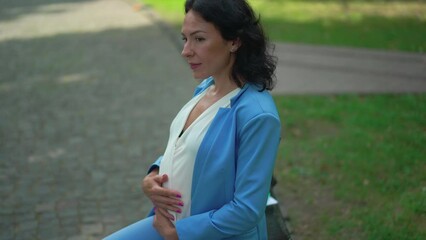 Side view successful pregnant businesswoman in elegant blue suit stroking belly dreaming sitting on bench in city. Happy confident Caucasian woman resting outdoors looking around. Slow motion - Powered by Adobe