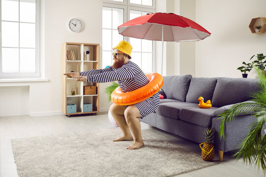 Funny chubby guy staying at home and having cool fake beach holiday in living room. Happy plus size man in hat and striped swimsuit pretends to jump in imaginary ocean waters. Summer vacation concept