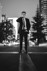 Obraz premium Young and attractive model guy with black suit on the middle of the street in the city downtown during sunset (in black and white)