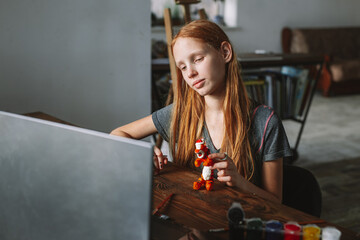 A beautiful cute red-haired girl creates crafts at home from a video lesson.. A cheerful teenager is engaged in a hobby on a laptop. Female student doing homework at homeschooling via video call.