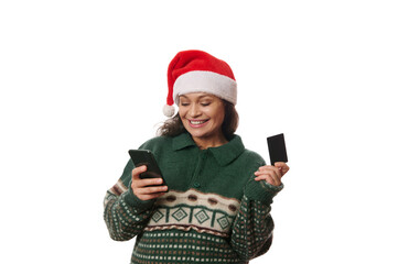 Cheerful Latin American woman wearing Xmas patterned sweater and Santa hat, holding credit card, making online payment while shopping in internet, using her smartphone, isolated over white background