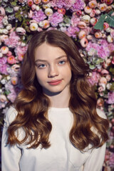 portrait of a teenage girl with long hair in a white sweater against a wall of flowers.