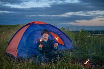 A teenage boy is happily relaxing in a tourist campsite, enjoying the view of the beautiful summer nature at sunset, waving sparklers.