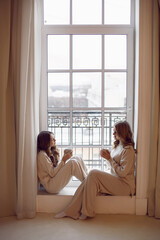 Obraz na płótnie Canvas Beautiful family mother and daughter in a white warm cozy suit are sitting next to the panoramic window with cups and drinking tea.