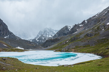 Atmospheric mountain landscape with frozen alpine lake and high snowy mountains. Awesome overcast scenery with icy mountain lake on background of snow mountains in low clouds. Scenic view to ice lake.