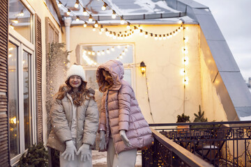 Obraz na płótnie Canvas family mother and daughter in jackets and a hat in winter are standing on the balcony on the street with garlands on Christmas day.