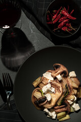 Plate of fried vegetables,fennel, mushrooms,chilli,cutlery,glass of wine. Black dishes on a dark background. Top view.