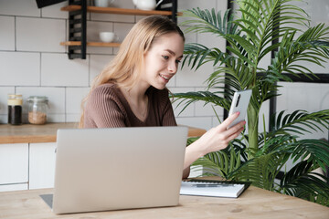 Beautiful smiling young female sitting at desk in kitchen using laptop computer and smartphone