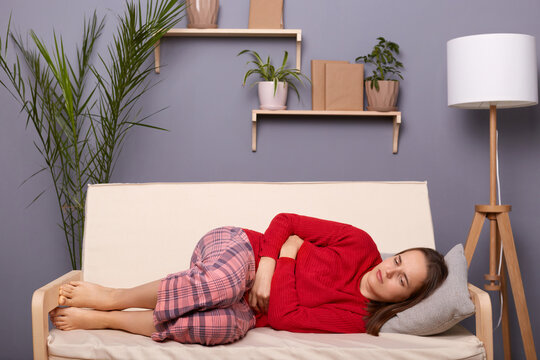 Full Length Portrait Of Unhealthy Sick Young Adult Woman With Dark Hair Wearing Red Sweater And Checkered Pants Lying On Sofa At Home, Suffering Terrible Stomachache, Feels Period Pain.