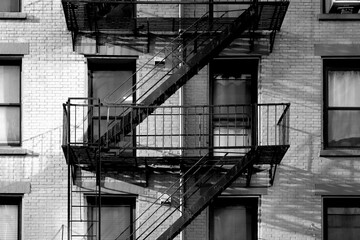 black and white close up New York City apartment building with brick exterior, windows, and diagonal fire escape stairs - obrazy, fototapety, plakaty