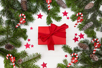 Green Christmas tree branches with pinecones decorated with red stars and Christmas candy canes, red gift centered on a white wooden background top view