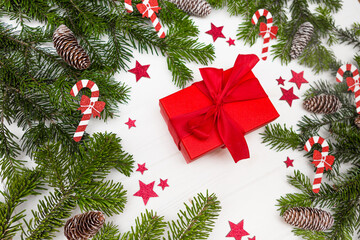 Green Christmas tree branches with pinecones decorated with red stars and Christmas candy canes, red gift centered on a white wooden background top view