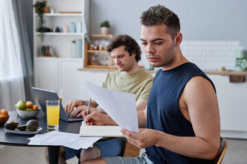 Portrait of young gay couple living together with focus on man studying at kitchen table