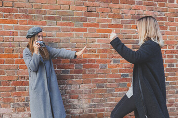 Girl takes picture of her female friend in front of brick wall in city street - photographer and vintage camera hobby concept