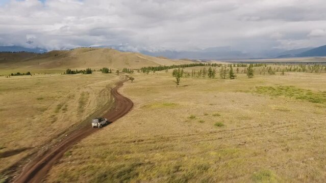 shooting with a copter from a height. A light-colored SUV drives along a winding road in a valley with sparse trees surrounded by high mountain ranges on a summer day.