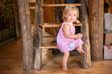 Toddler is sitting on a wooden ladder. Child in a pink dress, resting in a village house