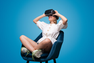 Young woman using glasses of virtual reality sitting on chair isolated over blue background....