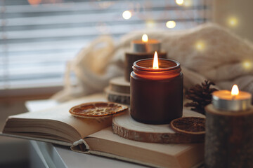 Cozy autumn or winter composition with aromatic candle, wool sweater, fairy lights, book. Aromatherapy, home atmosphere of cosiness and relax. Wooden background close up.