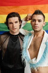 Vertical portrait of two gay young men posing with LGBTQ pride flag in background and wearing extravagant outfits