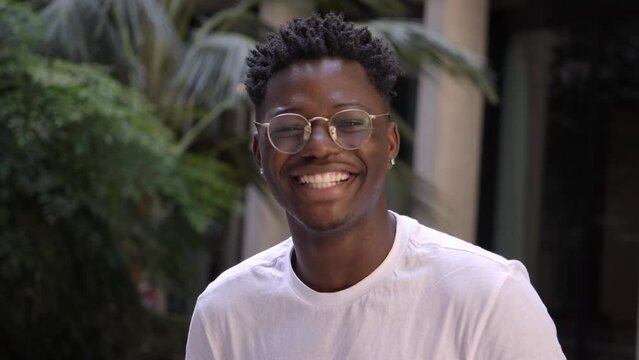 Portrait of black man very happy, smiling and looking at camera. An African American student sincerely smiles while standing outdoors. The guy is wearing a white t-shirt