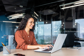 Happy and smiling hispanic businesswoman typing on laptop, office worker with curly hair and glasses happy with achievement results, at work inside office building. - obrazy, fototapety, plakaty
