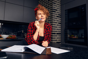 Joyful young Hispanic female working from home, doing paperwork, having coffee break, sitting at dining table with mug and diary, making to do list. Cute girl sketching in copybook and drinking tea