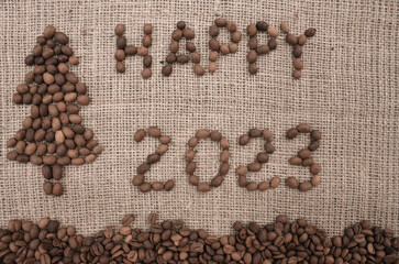 Merry Christmas and New Year greeting card. Text Happy 2023 in English and Christmas tree are made from coffee beans on fabric with burlap texture. View from above. New Year concept background