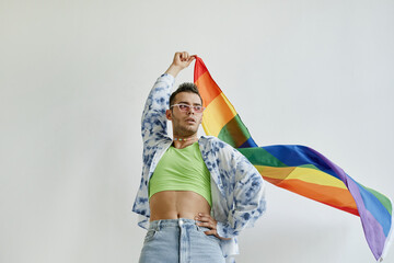 Low angle portrait of confident gay man waving pride flag against white background, copy space