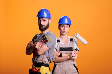 Professional renovators posing with painting tools, standing over yellow background. Team of constructors holding paintbrush and roller brush to work on refurbishment project in studio.