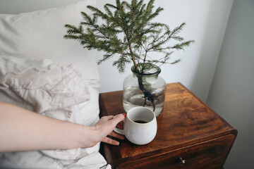 Close-up of womans hand holding cup of hot coffee, black tea drink. Morning breakfast in bedroom. Night table with little spruce tree. Cozy Christmas breakfast, lifestyle scene. Selective focus.