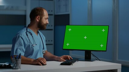 Medical assistant sitting at desk typing patient disease report on computer with green screen chroma key mock up display. Physician nurse working late at night in hospital office. Health care concept