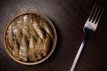 close-up photo of canned fish - sprats in oil and a shiny fork - on a dark wooden background - obrazy, fototapety, plakaty