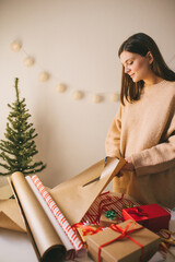 Young woman in knitted sweater wrapping Christmas presents using paper, scissors and colorful...
