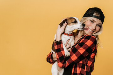 Adorable blonde in baseball cap and checked shirt , broad smiling, hugs her puppy Jack Russel, who...