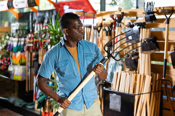 Obraz na płótnie Canvas Focused adult african american man looking for pitchfork to work in his garden at gardening supply store