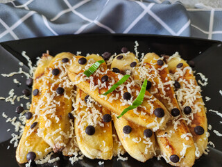 Grilled banana topped with grated cheese and chocolate chips. Served on black plate on grey textured background. Selective focus image.
