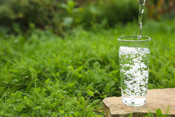 Pouring fresh water into glass on stone in green grass outdoors. Space for text