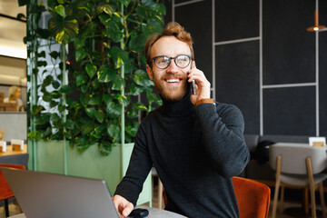 A young red-haired guy, a programmer or entrepreneur in glasses, sits in a stylish cafe at a laptop...