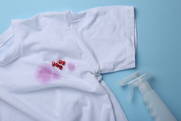 White shirt with fruit juicy stains, detergent and pomegranate seeds on light blue background, flat lay
