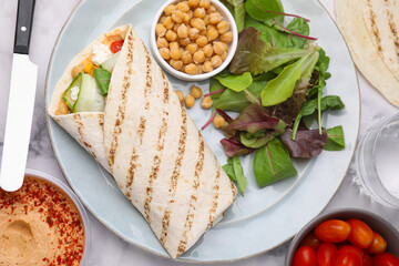 Plate with hummus wrap and vegetables on white marble table, flat lay