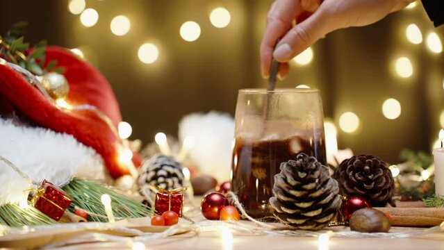 table with Christmas decorations and sweets, stirring hot chocolate with marshmallows. The garland bokeh is behind.