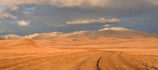 Winding ruts of field roads meet snow-covered mountain ranges through the autumn steppe on a warm evening.