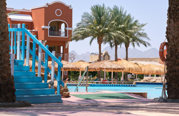 Empty deck chairs under straw shade umbrellas on swimming pool side in tropical resort. Summer vacations and getaway concept