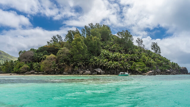 The small island is completely overgrown with tropical vegetation, palm trees. Boulders off the coast. The boat is moored near the shore in the turquoise ocean. Clouds in the blue sky. Seychelles.